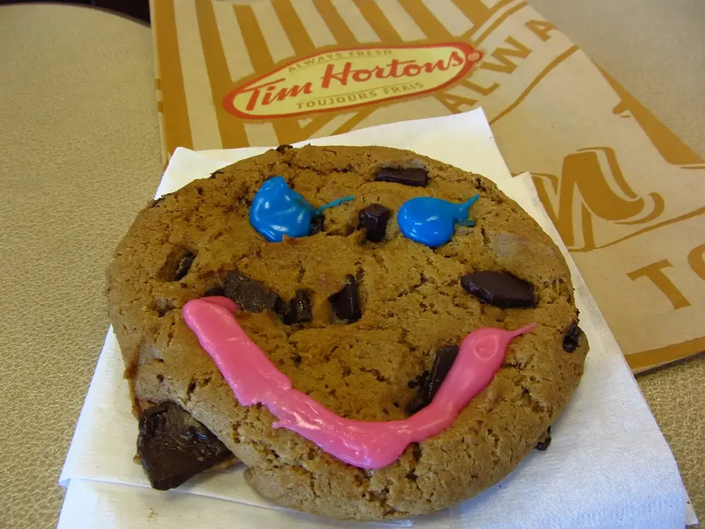 A Tim Hortons Smile Cookie on a napkin, with blue icing eyes, a pink icing smile and dark chocolate chunks